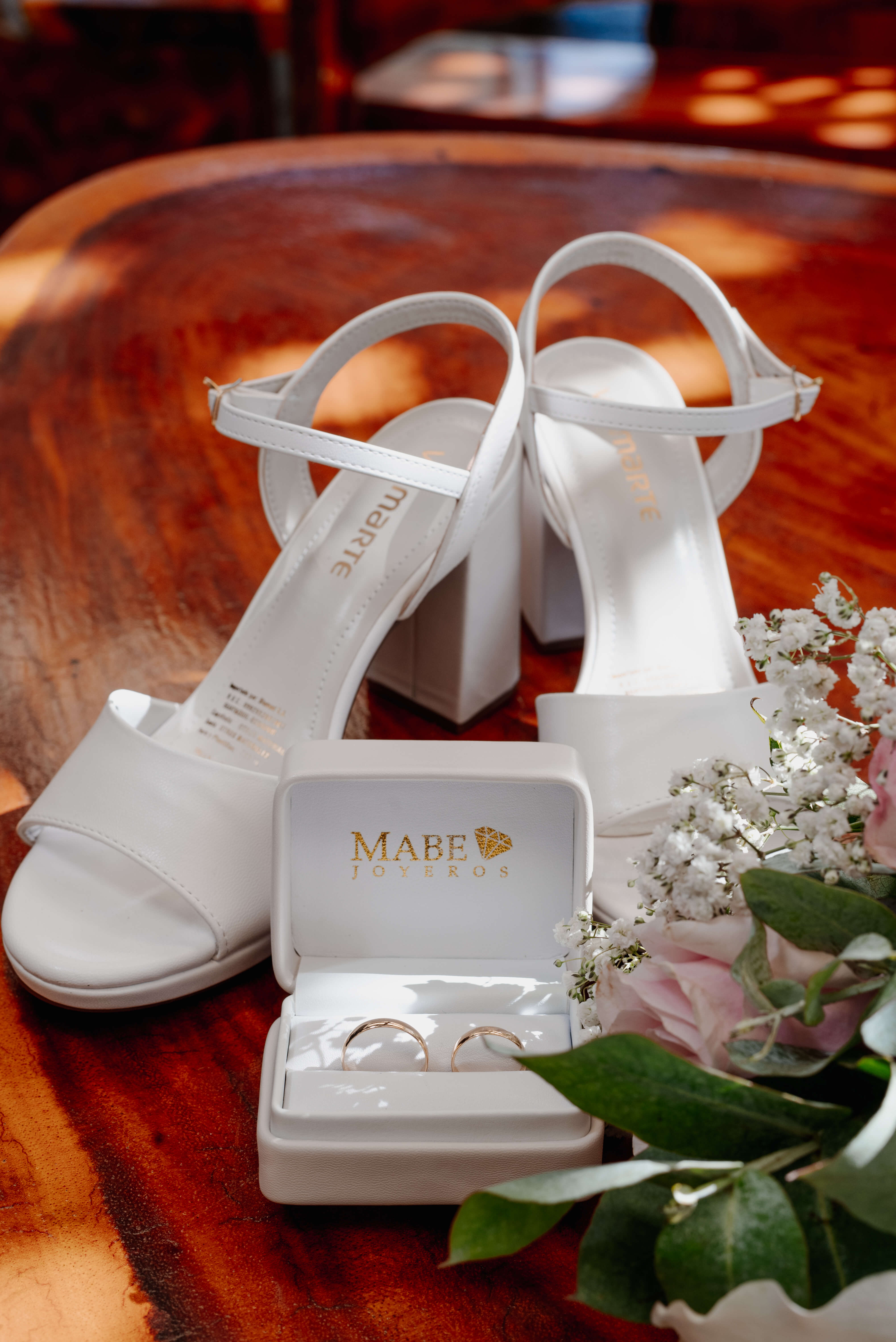 Zapatos blancos de novia, anillos en su estuche y flores sobre una mesa de madera.