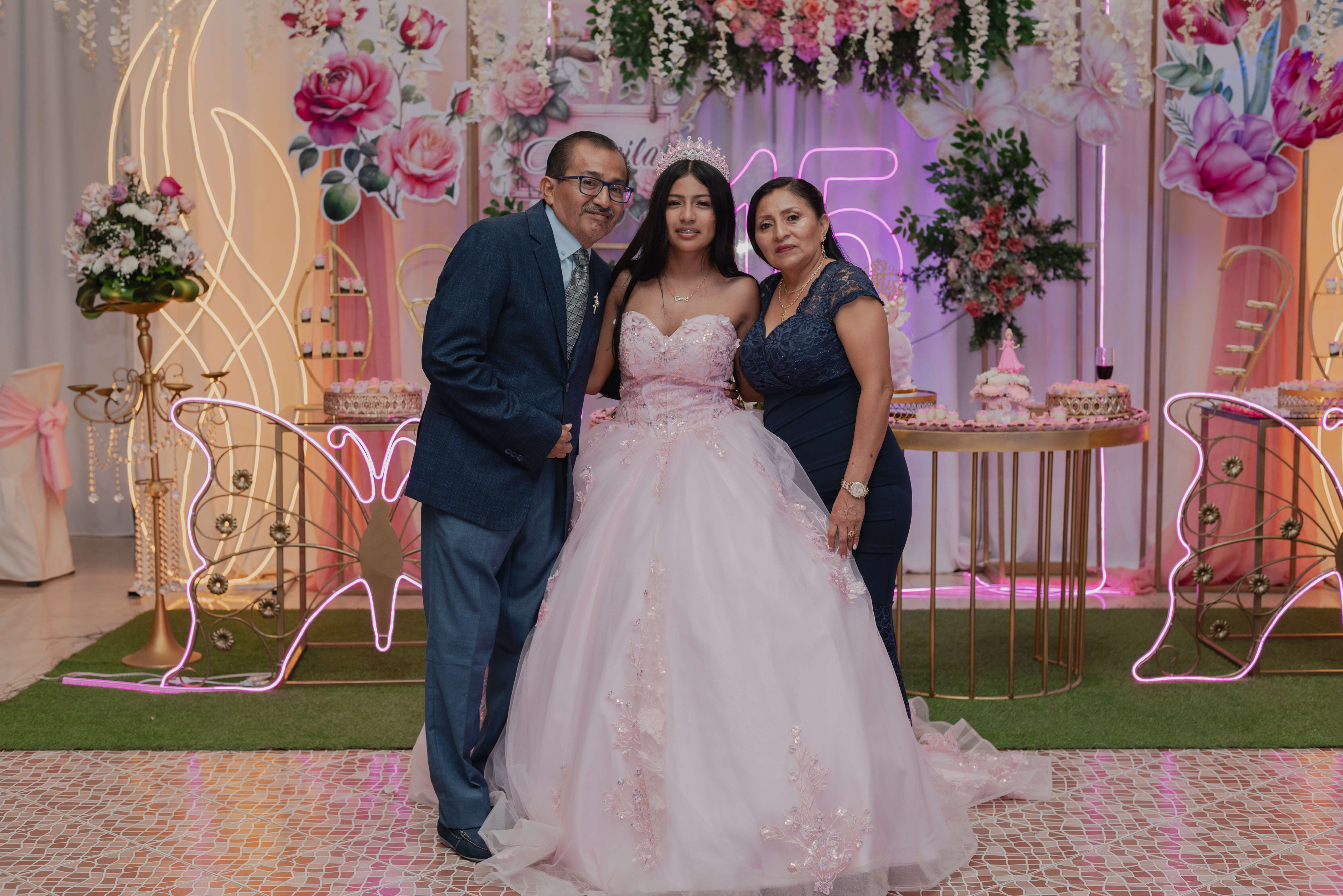 Camila sonriendo feliz mientras recibe un beso simultáneo en cada mejilla de parte de su padre y su madre dentro del salón.