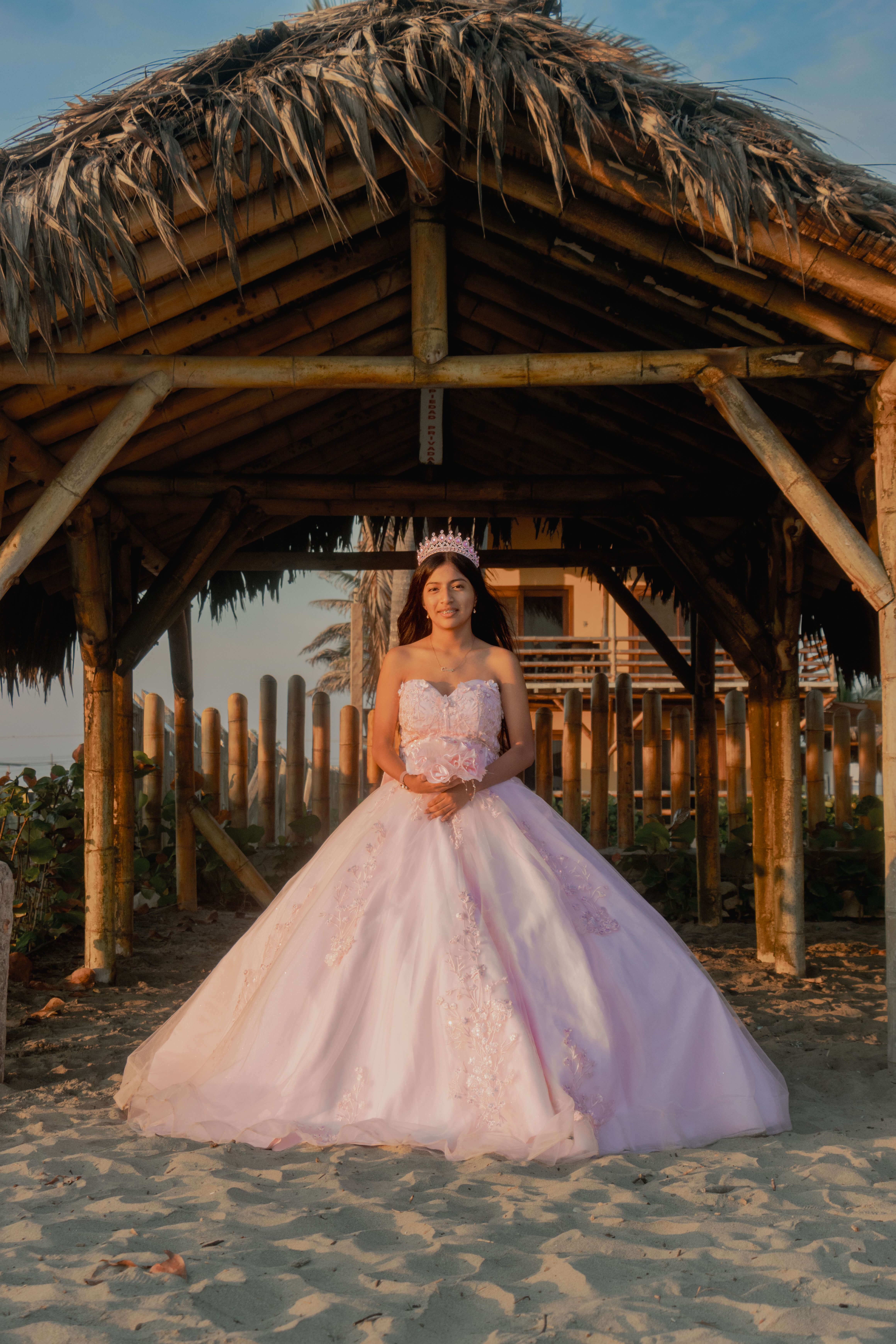 Quinceañera Camila sonriendo bajo una cabaña de paja en la playa vistiendo un vestido rosa de quince años.