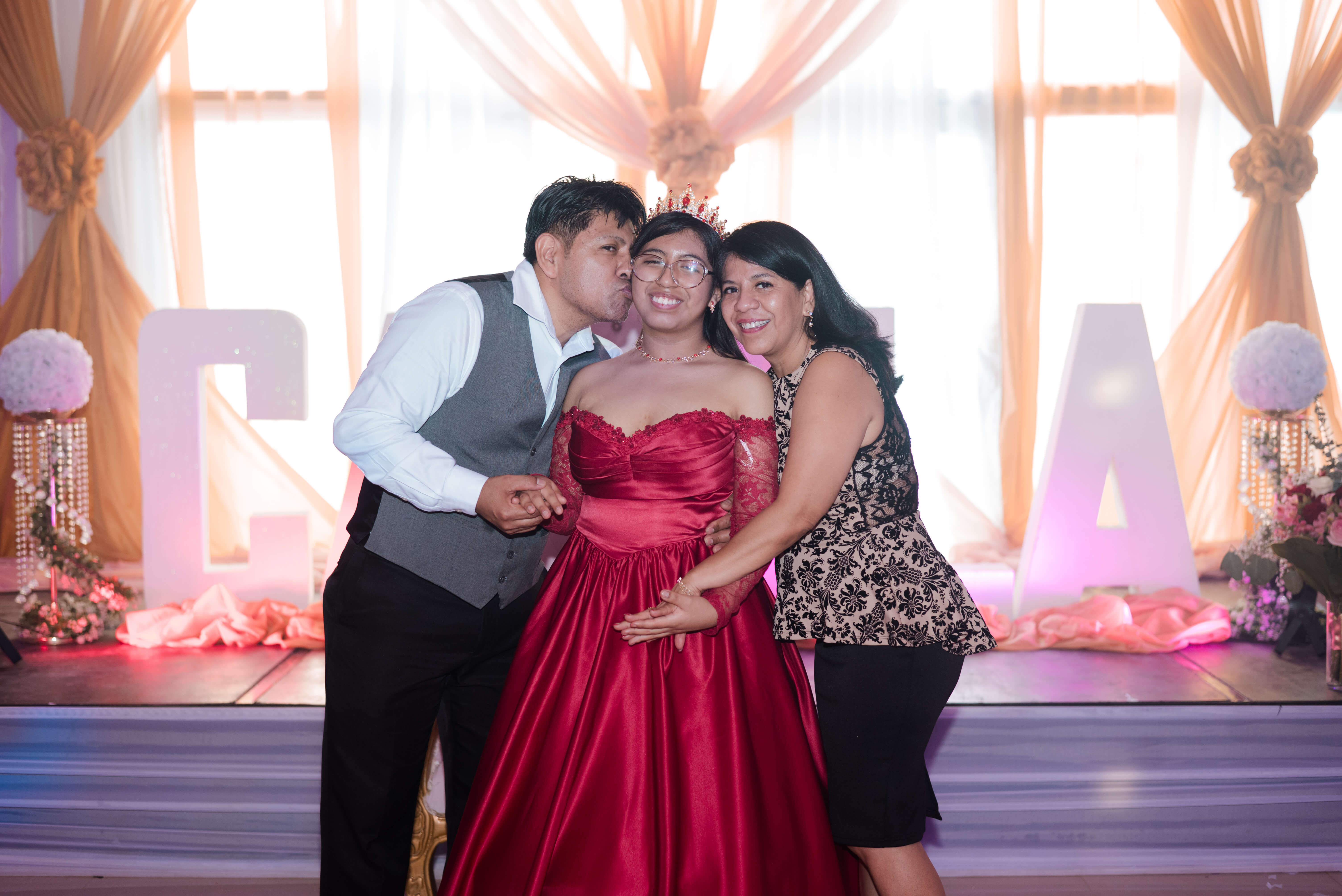 Los padres abrazados dándole un tierno beso en la mejilla a la quinceañera Camila.